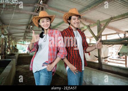 primo piano di una coppia di cowboy in piedi e sorridente i loro pollici in su in una stalla della fattoria della mucca Foto Stock