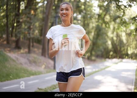 Donna ascoltare musica in esecuzione su asfalto pista estate parco caucasico Auricolari donne jogging mattina soleggiato Runner allenamento tenere smartphone in Healt. Mano Foto Stock