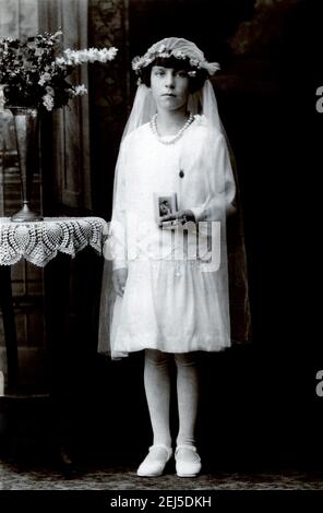 Foto d'archivio di una giovane ragazza che posa nel suo vestito bianco di comunione e velo bianco che tiene una carta religiosa, circa 1910. Foto Stock