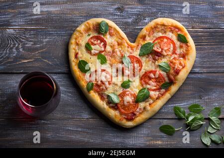 Deliziosa pizza margarita fatta in casa con pomodori e basilico a forma di cuore con un bicchiere di vino rosso su un tavolo di legno per San Valentino, top Foto Stock