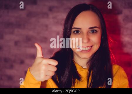 Ritratto di giovane donna sorridente e mostrante il pollice in su. Primo piano di una bruna felice che mostra un gesto di approvazione su sfondo retroilluminato. Foto Stock