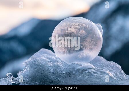 Belle bolle congelate seduti su un mucchio di neve e. ghiaccio con una delicata struttura di fiori di ghiaccio illuminati da il sole nascente con le montagne nel backgr Foto Stock