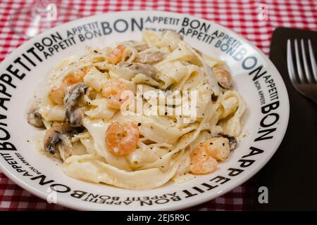 Gustose fettuccine con salsa di funghi e gamberi su un piatto che dice appetito Bon con tovaglia a scacchi rossi. Cucina italiana. Foto Stock