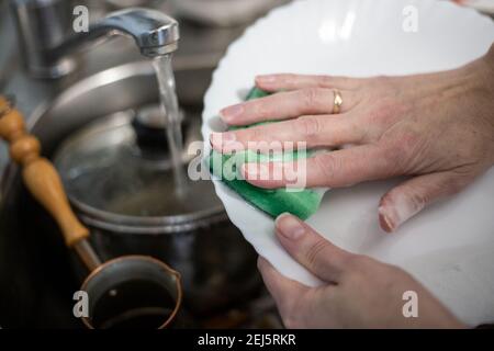 una donna lava i piatti con una spugna di schiuma, tiene un piatto nelle mani sopra il lavandino, l'acqua scorre dal rubinetto Foto Stock