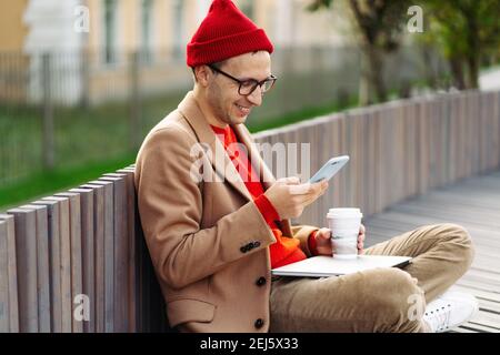 Uomo hipster che usa lo smartphone, chiacchierando nei social network, seduto all'aperto, bere caffè o tè. Foto Stock