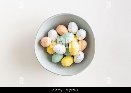 Una bella diffusione di cioccolato uovo di pasqua in colori pastello in una ciotola blu al centro su uno sfondo bianco, piatto con spazio copia Foto Stock