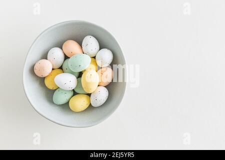 Una bella diffusione di cioccolato uovo di pasqua in colori pastello in una ciotola blu a sinistra su uno sfondo bianco, piatto con spazio copia Foto Stock