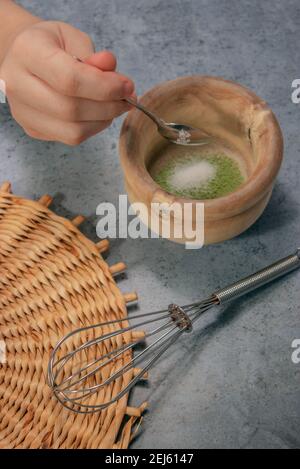 Immagine verticale della polvere di tè matcha e cucchiaio che versano lo zucchero nella ciotola di argilla sullo sfondo della pietra grigia. Preparazione del tè giapponese Matcha 2021. Foto Stock