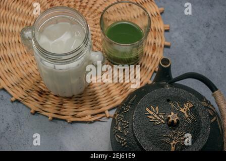 Immagine orizzontale vetro del latte ghiacciato liquido di tè verde formante e teiera su sfondo grigio pietra. Japaneses Green Tea Concept. Preparazione del tè Matcha 2021. Foto Stock