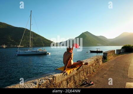 PERAST, MONTENEGRO - 14 LUGLIO 2016: Un uomo in costume da bagno sta prendendo il sole lungo la strada si sta proteggendo dalla luce del tramonto con un Foto Stock