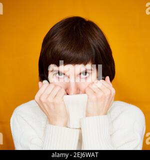 Nascondere la faccia dietro il collare del jumper avvolgerla in abiti caldi. Femmina di media età su parete gialla luminosa Foto Stock