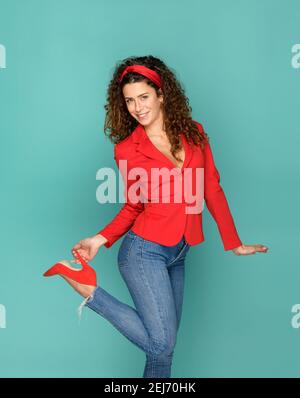 Donna sottile e positiva in un outfit elegante che sorride per la fotocamera e. tacco a contatto con la scarpa rossa su sfondo turchese Foto Stock