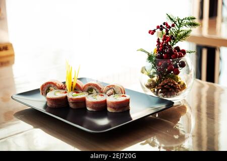 Sushi roll con pancetta e spigola fritta su un piatto nero, ingredienti spigola fritta, formaggio cremoso, pancetta, insalata Iceberg, uova di pesce volante, Salsa Teriyaki, riso, nori. Per il menu del ristorante Foto Stock