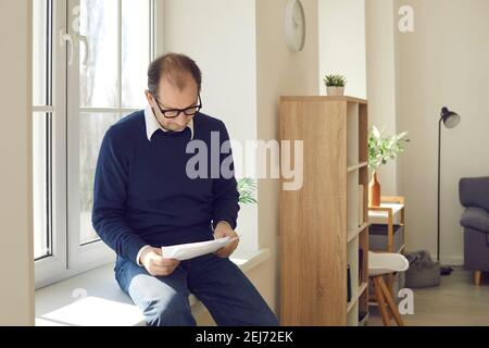 Uomo anziano che legge alcune lettere e documenti mentre si siede sopra davanzale a casa Foto Stock