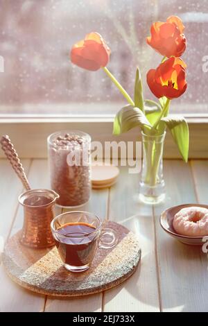 Caffè orientale in tradizionale caffettiera turca di rame con fiori sul davanzale della finestra. Davanzale in legno con tulipani arancioni e vaso di fiori di giacinto Foto Stock