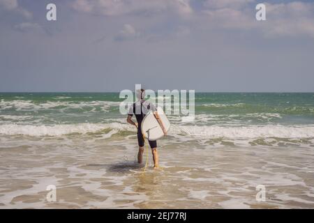 Giovane surfista in piedi contro l'oceano guardando le onde, surfista professionista in muta nera tenendo con una mano grande bella tavola da surf soleggiato Foto Stock