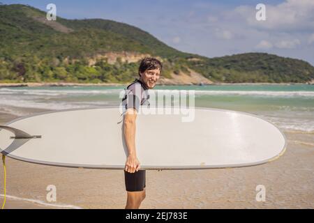 Giovane surfista in piedi contro l'oceano guardando le onde, surfista professionista in muta nera tenendo con una mano grande bella tavola da surf soleggiato Foto Stock