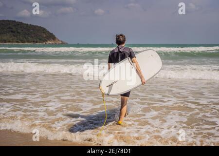 Giovane surfista in piedi contro l'oceano guardando le onde, surfista professionista in muta nera tenendo con una mano grande bella tavola da surf soleggiato Foto Stock