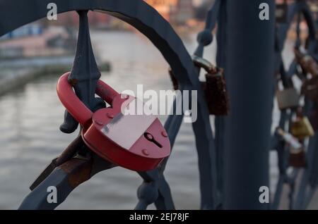 Lucchetto di ferro rosso chiuso su un elemento nero forgiato di recinzione ponte sul fiume sfocato. Su blocco attaccato barra dorata, su cui si può fare l'iscrizione. Co Foto Stock