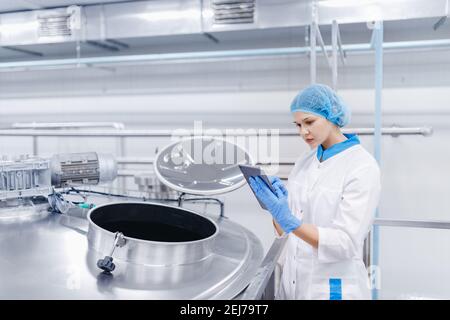 Produzione industriale moderna automatizzata alimentare. Operatrice donna controlla serbatoi d'acciaio con latte con computer tablet. Foto Stock