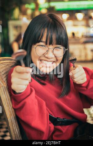 faccia divertente dell'adolescente asiatico solleva forchetta e cucchiaio sala da pranzo Foto Stock