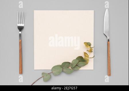 Coltello e forchetta con carta bianca per il testo del menu o della ricetta e ramo di eucalipto d'oro su tavolo grigio neutro. Mockup carta bianca e posate. Foto Stock