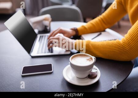 vista ritagliata della digitazione freelance sul computer portatile vicino a una tazza di caffè, sfondo sfocato Foto Stock