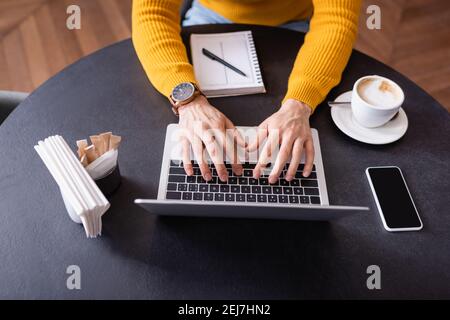 vista ritagliata della digitazione freelance sul laptop vicino a cup of caffè e notebook nel ristorante Foto Stock