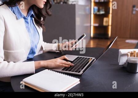 vista parziale del freelance che tiene lo smartphone mentre digita sul laptop in caffetteria Foto Stock