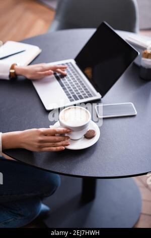 vista parziale della digitazione freelance sul computer portatile vicino allo smartphone e tazza di caffè nel ristorante, sfondo sfocato Foto Stock