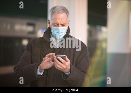 Uomo d'affari mascherato che utilizza il suo smartphone mentre attende il concetto di mobilità in treno, covid e coronavirus Foto Stock