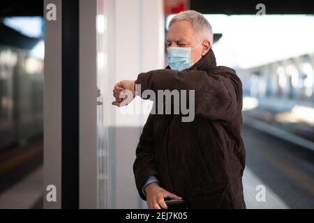 Uomo d'affari senior mascherato in attesa del concetto di mobilità del treno, del covid e del coronavirus Foto Stock