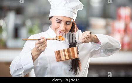 Una chef professionista puzza la salsa da un cucchiaio di legno che tiene una padella nell'altra mano. Foto Stock