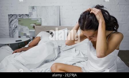 donna depressa che tocca la testa mentre si siede in camera da letto vicino a dormire uomo Foto Stock