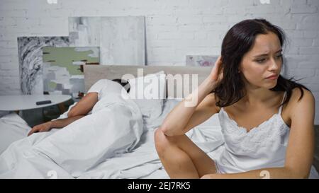 donna insoddisfatta che tocca i capelli mentre si siede sul letto vicino a dormire uomo Foto Stock