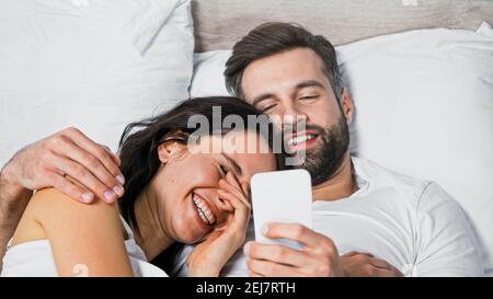 uomo sorridente utilizzando il telefono cellulare mentre abbracciando ragazza eccitato dentro letto Foto Stock