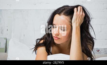scoraggiata giovane donna che tocca la testa mentre si siede in camera da letto Foto Stock