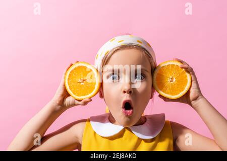 sorpresa ragazza in sciarpa tenendo le metà arancioni vicino alle orecchie isolate su rosa Foto Stock