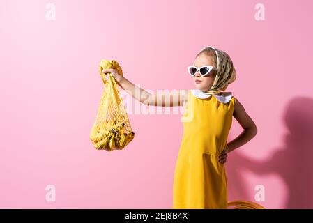 ragazza in copricapo e occhiali da sole che tengono una borsa a corda riutilizzabile con banane in piedi con la mano sull'anca in rosa Foto Stock