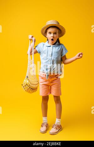 tutta la lunghezza della ragazza scioccata in cappello di paglia che tiene riutilizzabile borsa con le banane sul giallo Foto Stock