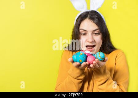 Una donna bruna felice con orecchie di coniglio sulla sua testa tiene uova di Pasqua e sorrisi. Preparatevi per Pasqua, su sfondo giallo con spazio di copia per il testo. Foto Stock