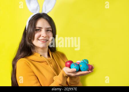 Una donna bruna felice con orecchie di coniglio sulla sua testa tiene uova di Pasqua e sorrisi. Preparatevi per Pasqua, su sfondo giallo con spazio di copia per il testo. Foto Stock