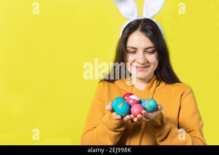 Una donna bruna felice con orecchie di coniglio sulla sua testa tiene uova di Pasqua e sorrisi. Preparatevi per Pasqua, su sfondo giallo con spazio di copia per il testo. Foto Stock