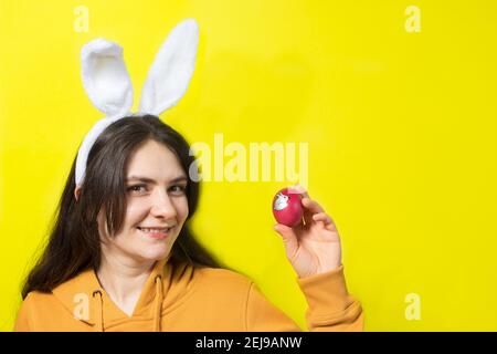 Una donna bruna felice con orecchie di coniglio sulla sua testa tiene uova di Pasqua e sorrisi. Preparatevi per Pasqua, su sfondo giallo con spazio di copia per il testo. Foto Stock