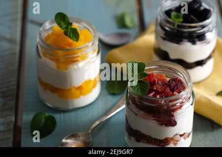 Yogurt bianco con arance di sangue, pezzi di pesca e more, con cucchiai, un tovagliolo giallo e foglie di menta, su uno sfondo di legno azzurro. Foto Stock