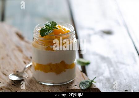 Yogurt bianco con pezzi di pesca e foglie di menta in un vasetto su una pietra gialla e sfondo di legno azzurro. Foto Stock