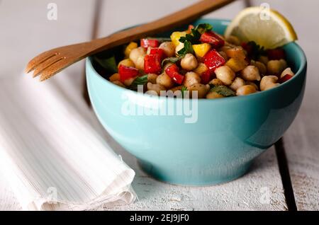 Insalata di ceci con peperoni rossi e gialli e prezzemolo in una ciotola turchese, con forchetta di legno e tovagliolo bianco, su sfondo bianco di legno. Foto Stock