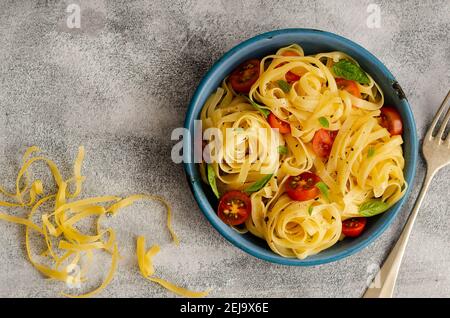 Fettuccine con pomodori ciliegini a fette e foglie di basilico in un piatto blu su sfondo grigio, con pasta cruda. Foto Stock