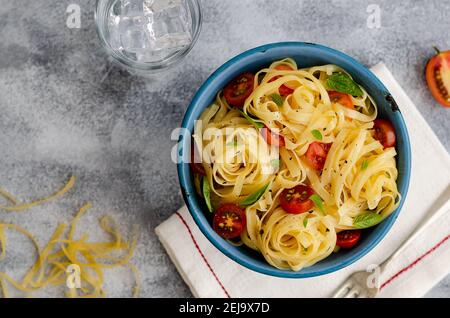 Fettuccine con pomodori ciliegini a fette e foglie di basilico in un piatto blu su sfondo grigio, con un tovagliolo bianco, forchetta, un bicchiere d'acqua e pasta cruda. Foto Stock