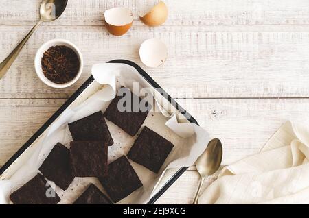 Brownie in un vassoio bianco con carta da forno, gusci d'uovo, cioccolato in una ciotola bianca, due cucchiai e un tovagliolo bianco, su uno sfondo di legno bianco. Foto Stock